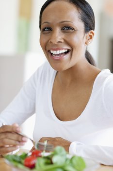 Photo of a woman looking at the camera and smiling. She is eating a green salad.